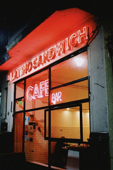 A nighttime scene features the entrance of a cafe bar with bright neon signs glowing in red that read 'LATINO SANDWICH' and 'CAFE BAR'. The interior, visible through the glass front, shows a warmly lit space with white-tiled walls and a person sitting at a table.