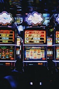 A row of colorful slot machines lit with bright neon lights, showcasing various graphical elements on the screens, indicating different games and jackpots. The atmosphere suggests a lively casino setting, bustling with excitement.