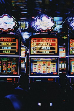 A row of colorful slot machines lit with bright neon lights, showcasing various graphical elements on the screens, indicating different games and jackpots. The atmosphere suggests a lively casino setting, bustling with excitement.