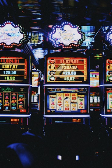 A row of colorful slot machines lit with bright neon lights, showcasing various graphical elements on the screens, indicating different games and jackpots. The atmosphere suggests a lively casino setting, bustling with excitement.