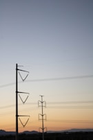 Power lines stretch across the sky supported by two tall transmission towers. The sky transitions from a soft blue at the top to a warm orange near the horizon, suggesting either sunrise or sunset. The landscape below is mostly flat with silhouetted hills in the distance.