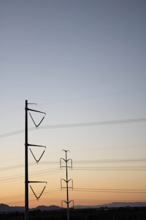 Power lines stretch across the sky supported by two tall transmission towers. The sky transitions from a soft blue at the top to a warm orange near the horizon, suggesting either sunrise or sunset. The landscape below is mostly flat with silhouetted hills in the distance.