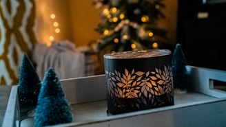 A dimly lit scene featuring a decorative candle with leaf patterns on a tray. Miniature Christmas trees are placed around the candle, and in the background, blurred holiday lights and a Christmas tree are visible, creating a cozy atmosphere.