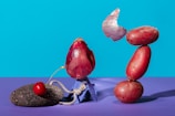 An abstract composition with an onion perched on a small blue stand next to a pumice stone holding a cherry, connected by a piece of rope. On the right, three red potatoes are precariously stacked, with an onion peel floating above them. The background is a solid bright blue, and the surface is a contrasting purple.