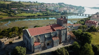 A historic stone building with a red-tiled roof surrounded by lush greenery, located near a serene river with a bridge in the background. The landscape features a charming town with several houses nestled on rolling hills.