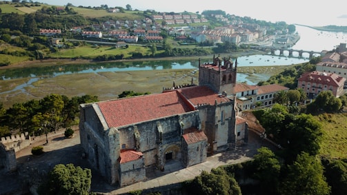 A historic stone building with a red-tiled roof surrounded by lush greenery, located near a serene river with a bridge in the background. The landscape features a charming town with several houses nestled on rolling hills.