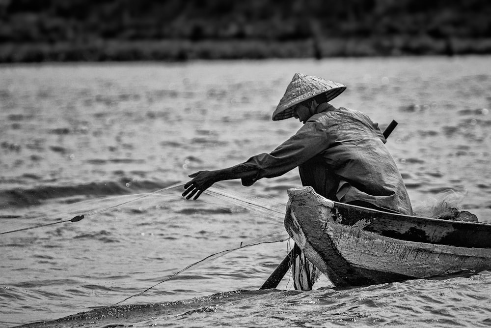 grayscale photo of man in hat and long sleeve shirt sitting on boat on water