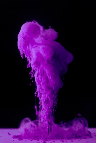Vibrant purple ink or pigment is swirling and billowing upward in a dramatic plume against a dark background, with fluid motion and soft, cloud-like textures.