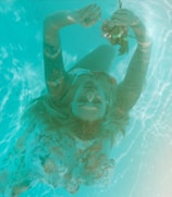 A person is submerged in water, holding flowers, with eyes closed and a peaceful expression. The image has a dreamlike, serene quality, with the sunlight filtering through the water creating a pattern on the person's skin.