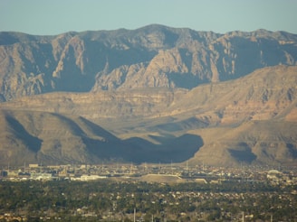 A vast desert landscape with rugged mountain ranges in the background and a small town or residential area nestled in the valley below. The terrain transitions from the greenery of the town to the barren, rocky mountains.