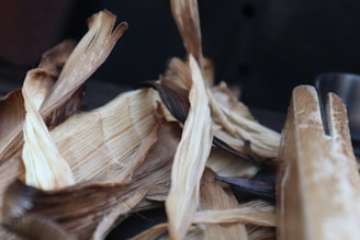 A close-up view of dried corn husks with varying shades of brown and beige, lying on a dark surface. The texture of the husks is rough and fibrous, and the edges are slightly curled.