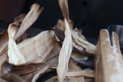 A close-up view of dried corn husks with varying shades of brown and beige, lying on a dark surface. The texture of the husks is rough and fibrous, and the edges are slightly curled.