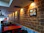 A restaurant interior with a brick wall featuring posters and warm ambient lighting. The tables and chairs are arranged along the wall, with a few decorative items hanging from the ceiling. The corridor extends towards an open area with visible outdoor light.