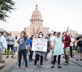 A group of people gather on a street near a historic building, with one person holding a sign that reads 'Jesus Loves You.' Several individuals have their arms raised, and the scene appears to be a peaceful demonstration or gathering.