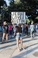 A woman stands in a public area holding a sign that reads 'Jesus Loves Austin.' She is surrounded by a group of people who appear to be gathered for an outdoor event. The setting is in a park-like area with trees and paved walkways. The woman is wearing casual clothing, including a black shirt and denim shorts.