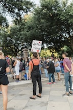 A group of people gather outdoors, surrounded by trees and standing on a paved area. One person in the foreground is holding a sign with the text 'Jésus t'aime' written on it, decorated with red hearts. The crowd appears to be engaged in some form of public event or demonstration.