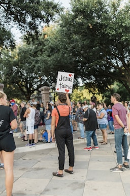 A group of people gather outdoors, surrounded by trees and standing on a paved area. One person in the foreground is holding a sign with the text 'Jésus t'aime' written on it, decorated with red hearts. The crowd appears to be engaged in some form of public event or demonstration.