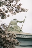 A stone statue of a muscular figure holding a trident sits atop a building, partially obscured by branches of a blooming tree. The sculpture presents a sense of strength and classical beauty, enhanced by the soft focus of the surrounding pink blossoms.