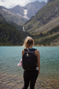 A person with blonde hair is standing at the edge of a serene turquoise lake, surrounded by tall, rugged mountains with patches of greenery. The individual is wearing a black backpack adorned with small trinkets and appears to be in a contemplative stance, gazing at the natural scenery.