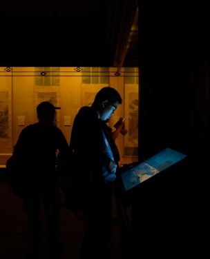 In a dimly lit room, two silhouetted figures stand near an interactive display screen emitting a pale blue light. The background is illuminated by a warm, yellowish glow coming from framed displays on the wall.