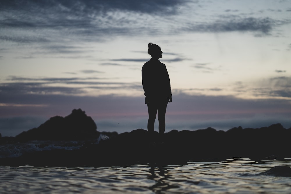 silhouette of man standing on rock formation near body of water during sunset