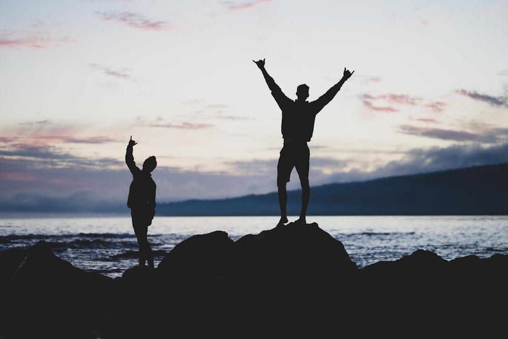 silhouette of 2 women standing on rock formation near body of water during sunset