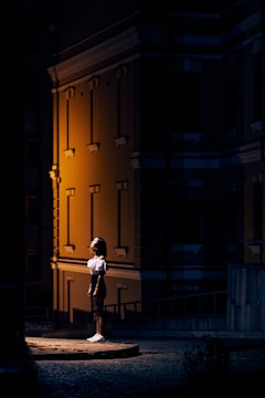A person is standing on a platform in a dimly lit urban environment, illuminated by a warm light from the side. The backdrop features a building with architectural details, casting shadows that contrast with the light.