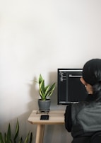 A person with long hair sits at a desk, facing a computer monitor displaying code or text in a dark-themed editor. The workspace includes a small table with a potted snake plant, a pair of glasses on a stand, and a smartphone placed on the table surface.
