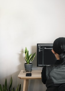 A person with long hair sits at a desk, facing a computer monitor displaying code or text in a dark-themed editor. The workspace includes a small table with a potted snake plant, a pair of glasses on a stand, and a smartphone placed on the table surface.