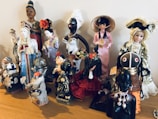 A collection of diverse, cultural figurines arranged on a wooden surface. The dolls are dressed in traditional attire from various cultures, including elaborate dresses, headpieces, and accessories. Materials vary, with some made of ceramic, fabric, or wood, showcasing intricate details and craftsmanship.