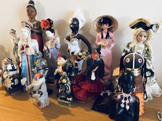 A collection of diverse, cultural figurines arranged on a wooden surface. The dolls are dressed in traditional attire from various cultures, including elaborate dresses, headpieces, and accessories. Materials vary, with some made of ceramic, fabric, or wood, showcasing intricate details and craftsmanship.