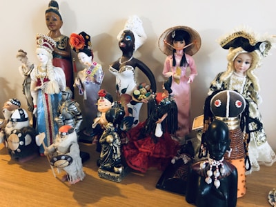 A collection of diverse, cultural figurines arranged on a wooden surface. The dolls are dressed in traditional attire from various cultures, including elaborate dresses, headpieces, and accessories. Materials vary, with some made of ceramic, fabric, or wood, showcasing intricate details and craftsmanship.