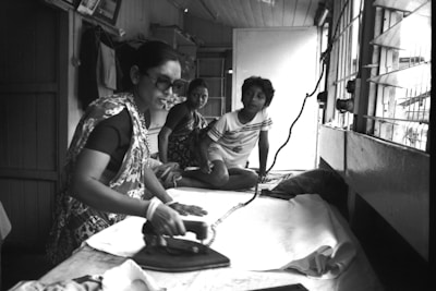 A woman is using an iron on a white fabric inside a modest room, with two children sitting nearby. The interior is simple with wooden walls, a window covered with bars, and hanging clothes. The atmosphere suggests a home setting or a small tailoring shop.