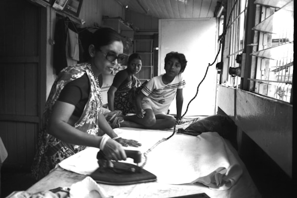 A woman is using an iron on a white fabric inside a modest room, with two children sitting nearby. The interior is simple with wooden walls, a window covered with bars, and hanging clothes. The atmosphere suggests a home setting or a small tailoring shop.