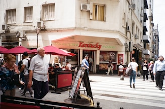 A street scene in an urban area featuring a corner pizzeria with a sign reading 'Kentucky Pizzeria' and several people walking and sitting at tables under red umbrellas. The building has a beige facade with multiple windows and air conditioning units. Pedestrians are crossing a zebra-striped crosswalk.