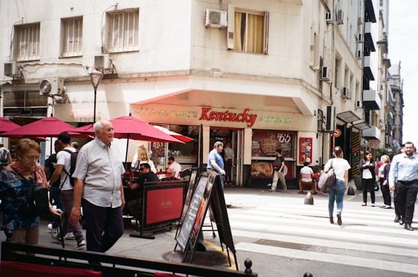 A street scene in an urban area featuring a corner pizzeria with a sign reading 'Kentucky Pizzeria' and several people walking and sitting at tables under red umbrellas. The building has a beige facade with multiple windows and air conditioning units. Pedestrians are crossing a zebra-striped crosswalk.