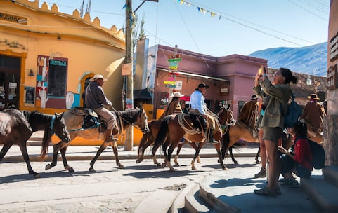 A group of people on horseback is riding through a sunlit street in a vibrant town. Colorful buildings and signs are visible in the background, with one person standing nearby taking a photograph. The scene captures a lively, cultural atmosphere.