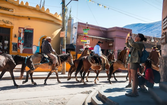 A group of people on horseback is riding through a sunlit street in a vibrant town. Colorful buildings and signs are visible in the background, with one person standing nearby taking a photograph. The scene captures a lively, cultural atmosphere.