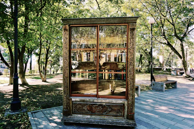 An ornate wooden cabinet is placed outdoors, resembling a public bookshelf, and is filled with various books. The cabinet features intricate carvings and appears to stand on a paved path in a park setting. Surrounding it are green trees, park benches, and a lamp post, creating a peaceful and inviting atmosphere.