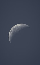 A detailed view of the moon in a crescent phase, against a dark, clear sky. The lunar surface reveals visible craters and textures emphasizing its rugged terrain.