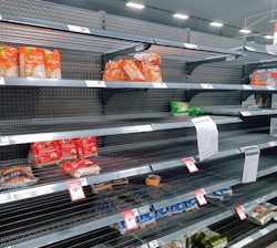 Nearly empty supermarket shelves with a few remaining packs of rice and other food items. Price tags are visible, and there is a printed notice attached to one of the shelves.
