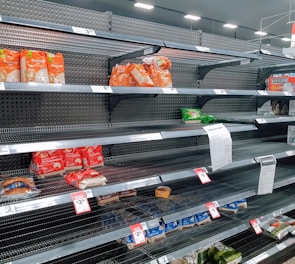 Nearly empty supermarket shelves with a few remaining packs of rice and other food items. Price tags are visible, and there is a printed notice attached to one of the shelves.