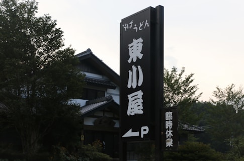 A large vertical sign with Japanese characters stands in front of a traditional building with a tiled roof. The surrounding area has lush green trees and foliage.