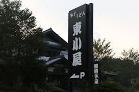 A large vertical sign with Japanese characters stands in front of a traditional building with a tiled roof. The surrounding area has lush green trees and foliage.