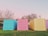 Three brightly colored shipping containers are placed side by side on lush green grass. The left container is painted blue, the center one is pink, and the right is yellow. Behind the containers, leafless trees stand under a pastel pink and blue sky, suggesting a serene and calm environment.