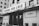 A black and white image featuring the facade of a cinema named Cinema São Luiz. The building appears old with some weathering visible. The marquee displays the text 'XII Janela'. Several people are gathered at the entrance, and a few cars are parked in the foreground.