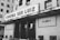 A black and white image featuring the facade of a cinema named Cinema São Luiz. The building appears old with some weathering visible. The marquee displays the text 'XII Janela'. Several people are gathered at the entrance, and a few cars are parked in the foreground.
