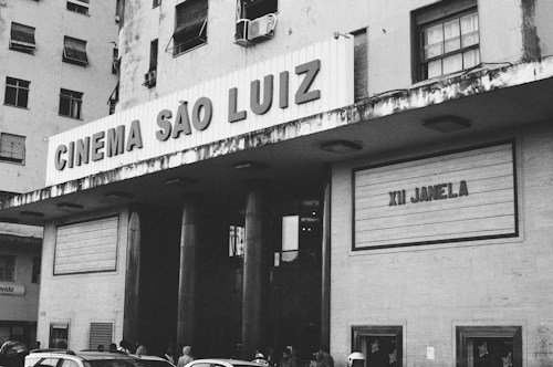 A black and white image featuring the facade of a cinema named Cinema S&atilde;o Luiz. The building appears old with some weathering visible. The marquee displays the text 'XII Janela'. Several people are gathered at the entrance, and a few cars are parked in the foreground.