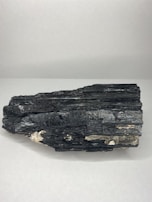 A large black mineral sample with a layered structure and shiny surface, resting on a plain white background. The rock has some lighter brown and white inclusions.