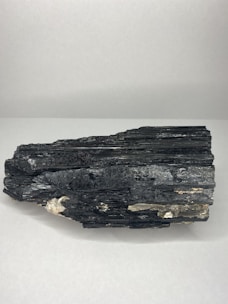 A large black mineral sample with a layered structure and shiny surface, resting on a plain white background. The rock has some lighter brown and white inclusions.